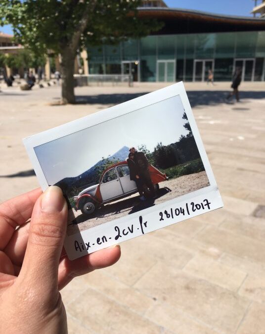 Main tenant un Polaroid de la 2CV d’Aix en 2CV devant une place aixoise